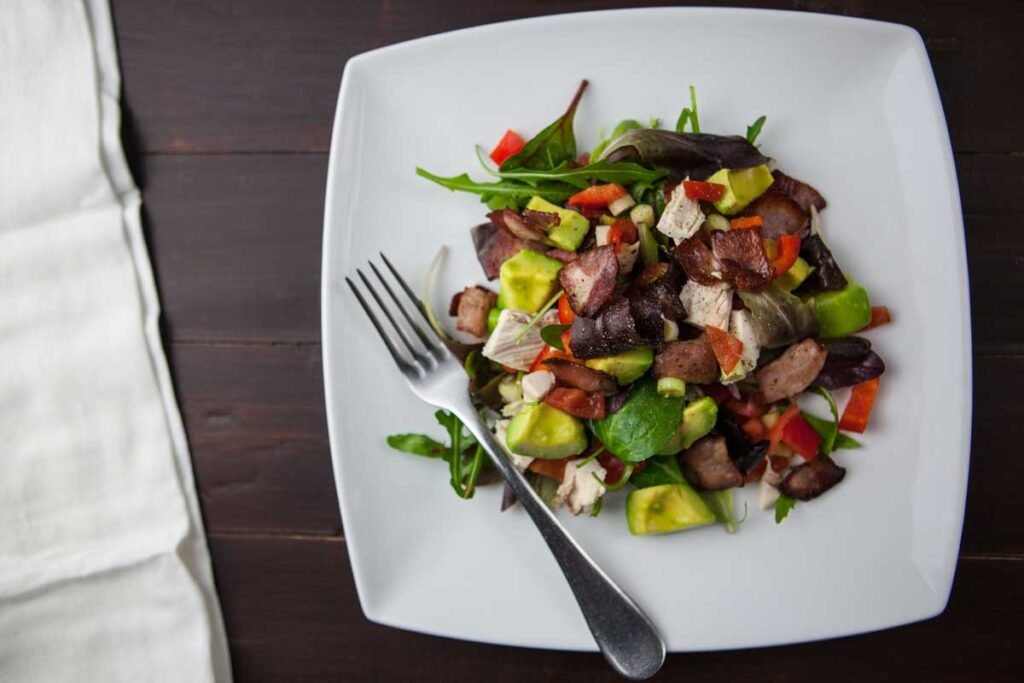 Top-down view of a white square plate with a colorful salad—avocado, greens, chicken, and diced red peppers—plus a fork resting on the plate.