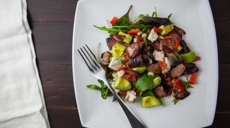 Top-down view of a white square plate with a colorful salad—avocado, greens, chicken, and diced red peppers—plus a fork resting on the plate.