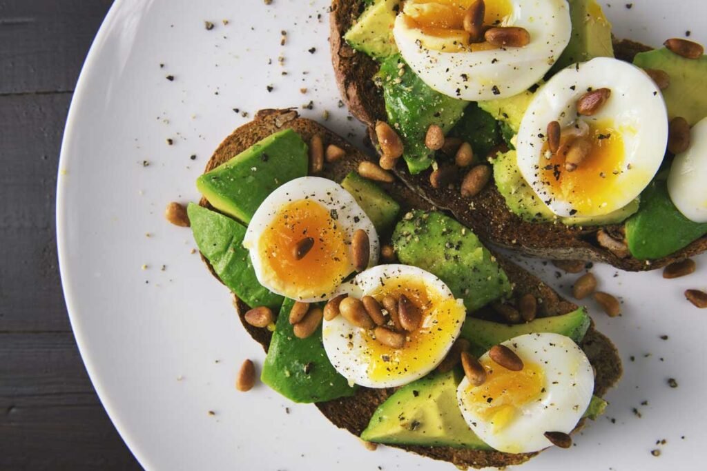Toast topped with avocado, sliced hard-boiled eggs, and pine nuts on dark-grain bread, sprinkled with pepper on a white plate.