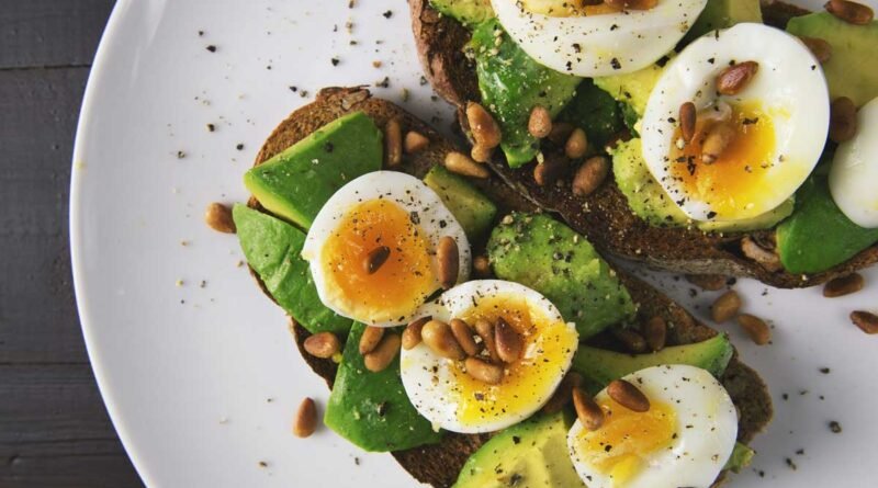 Toast topped with avocado, sliced hard-boiled eggs, and pine nuts on dark-grain bread, sprinkled with pepper on a white plate.