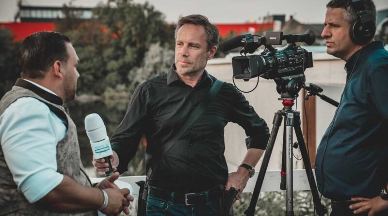 A reporter holds a microphone toward a man in a black shirt while two camera operators film on a rooftop.