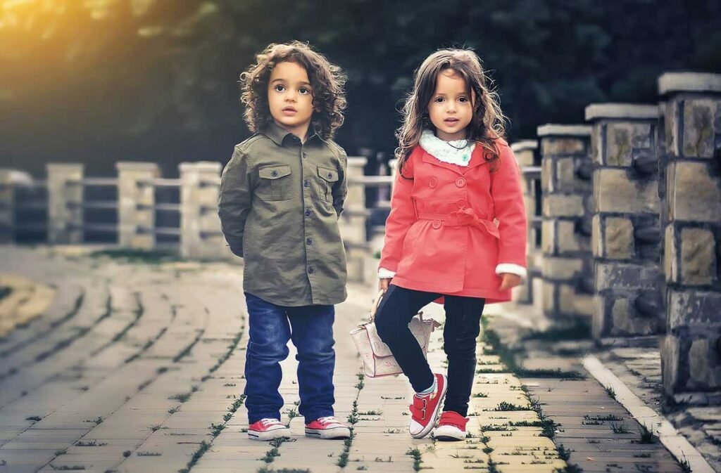 Two young children stand on a cobblestone path outdoors, the girl in a pink coat and the boy in an olive jacket.