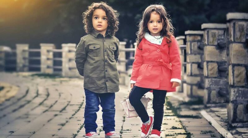 Two young children stand on a cobblestone path outdoors, the girl in a pink coat and the boy in an olive jacket.