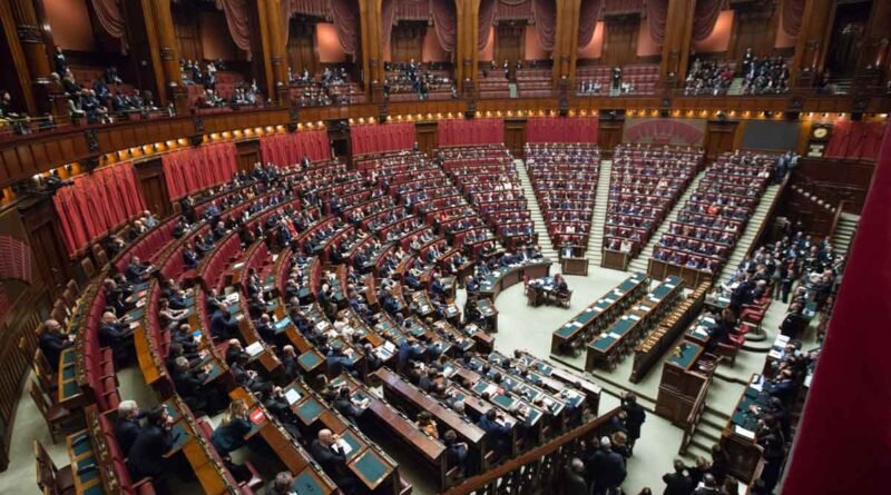A grand parliamentary chamber with semicircular rows of red seats, wood paneling, and a central desk area with officials gathered.