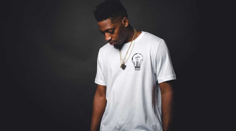 Man in a white T-shirt with a lightbulb graphic, gold chains, looking down against a dark studio background.