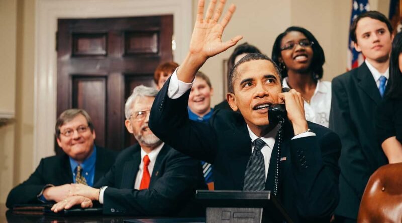 A man in a dark suit sits at a desk, speaking on a phone and raising his hand in a wave as a group of people watches behind him.