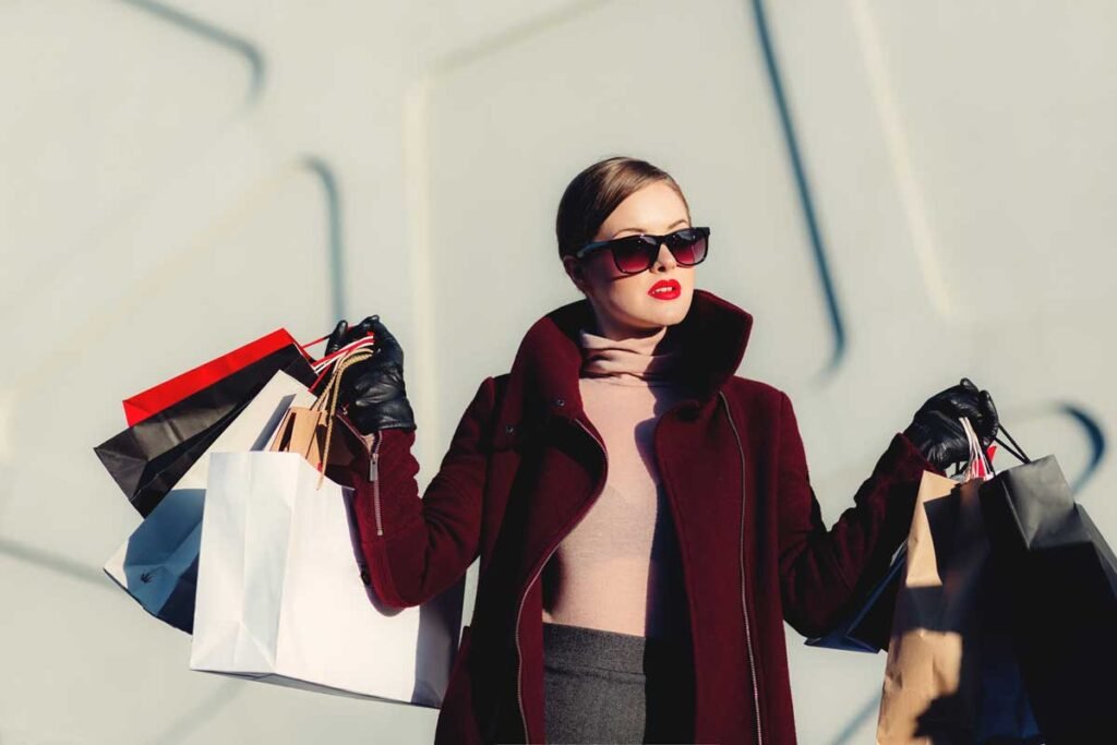 Stylish woman in a burgundy coat and oversized sunglasses holds multiple shopping bags outside.
