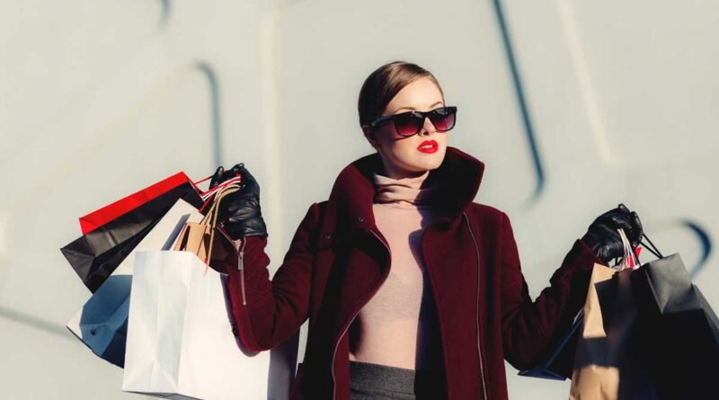 Stylish woman in a burgundy coat and oversized sunglasses holds multiple shopping bags outside.
