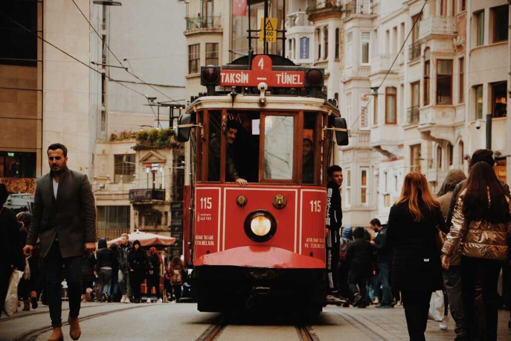 Front view of a red tram moving along tracks in a busy city street with pedestrians on both sides.