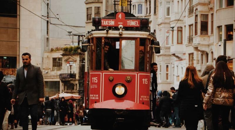 Front view of a red tram moving along tracks in a busy city street with pedestrians on both sides.