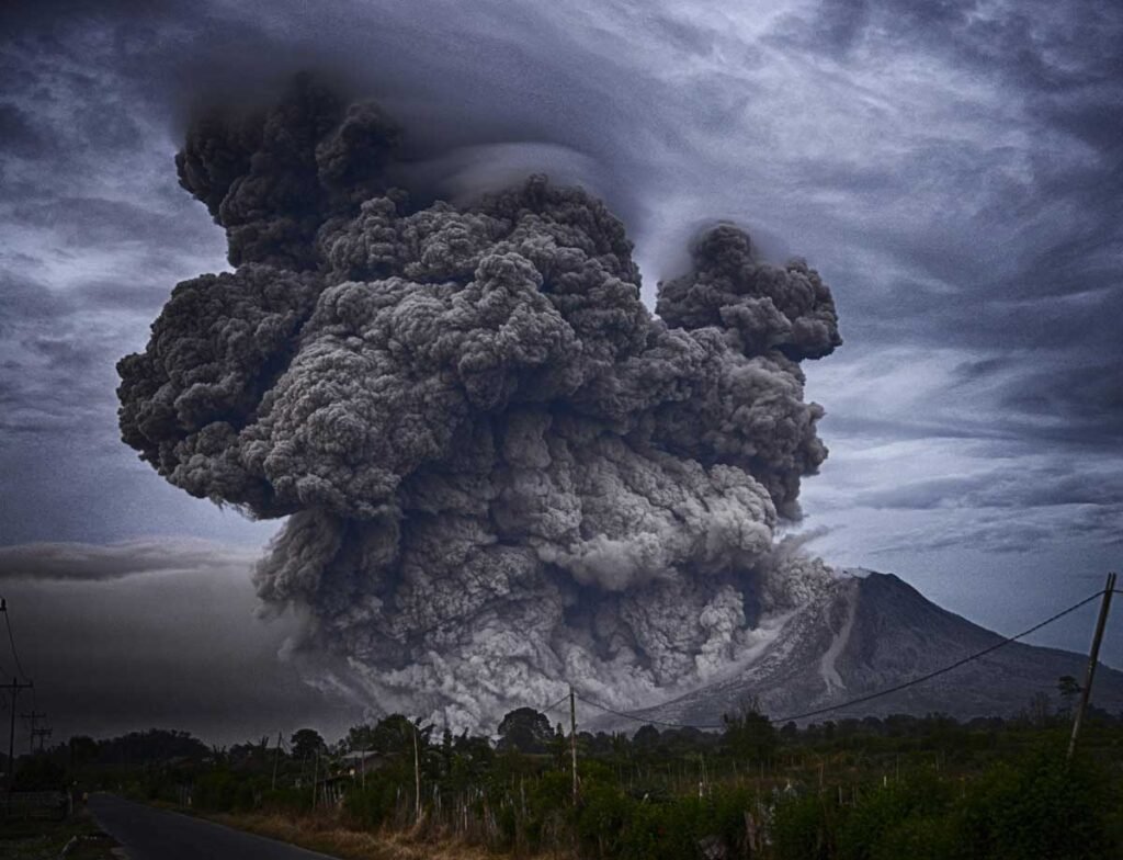 Volcanic eruption spewing a massive dark ash cloud over a rural landscape with a mountain in the background and power lines in the foreground.